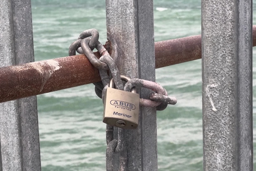 A padlock and chain secures a silver gate to a rusted metal handrail on a jetty. 
