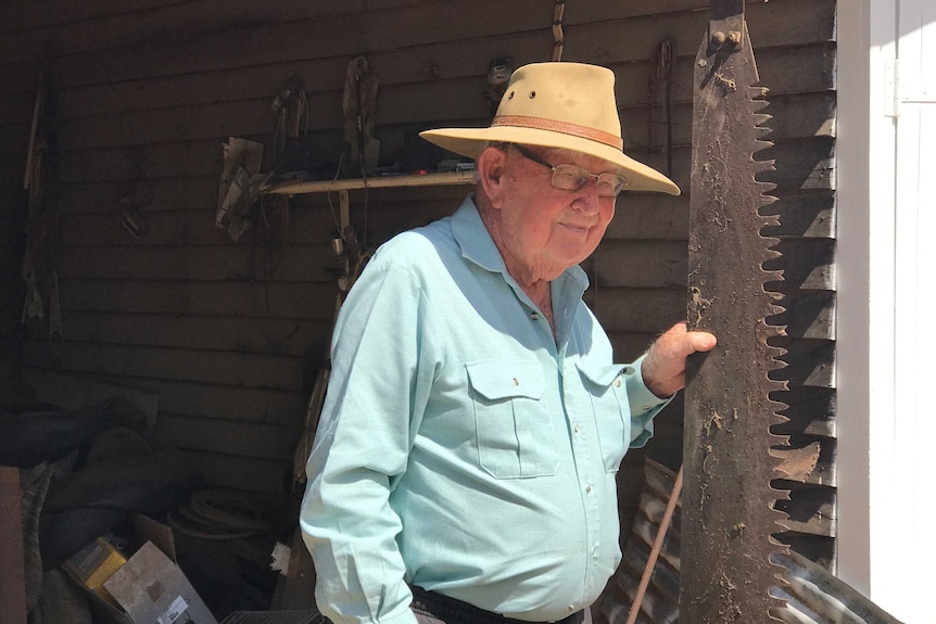 Cattle farmer Max Hockey stands out the front of a shed while holding a vintage saw.