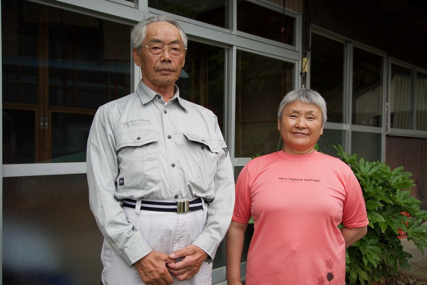An older Japanese couple stand in front of a house