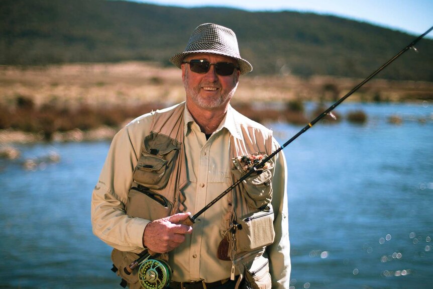 Col Sinclair looks to the camera and smiles while wearing glasses and fishing gear and holding a fishing rod.