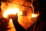 Man's face lit by fire as he pulls a brick out of a kiln and fire bursts out.
