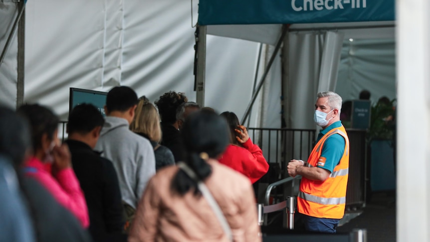 A group of people queue watched by an official in a hi-vis vest