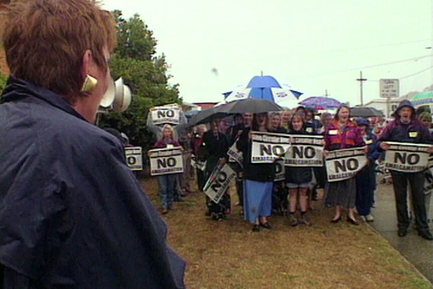 Men and women stand holding up 'No' signs in the rain, some with umbrellas as a woman shouts 'No' into a microphone.