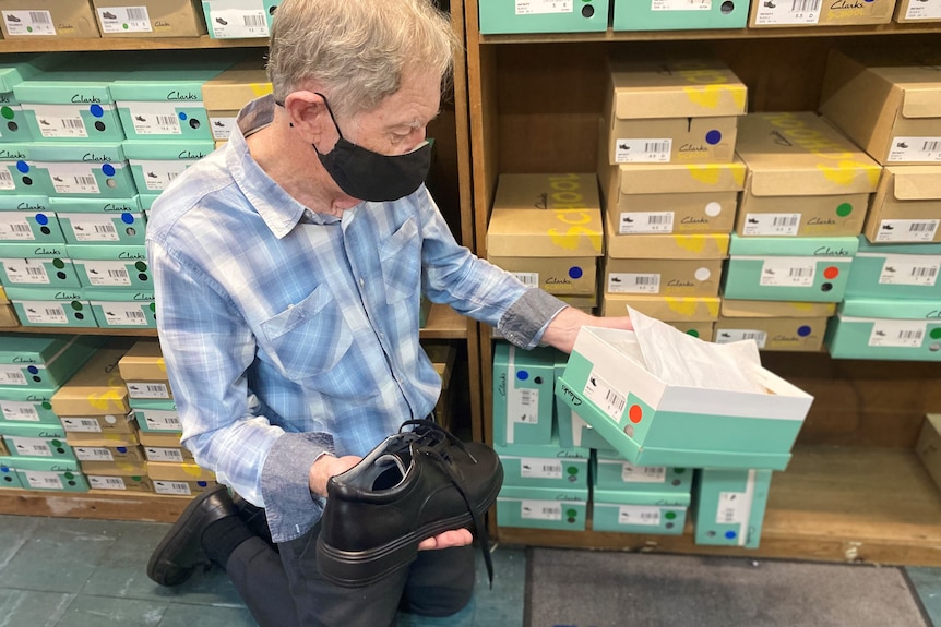 A man kneels in front of a shelf filled with shoe boxes, holding an open box and a black shoe.