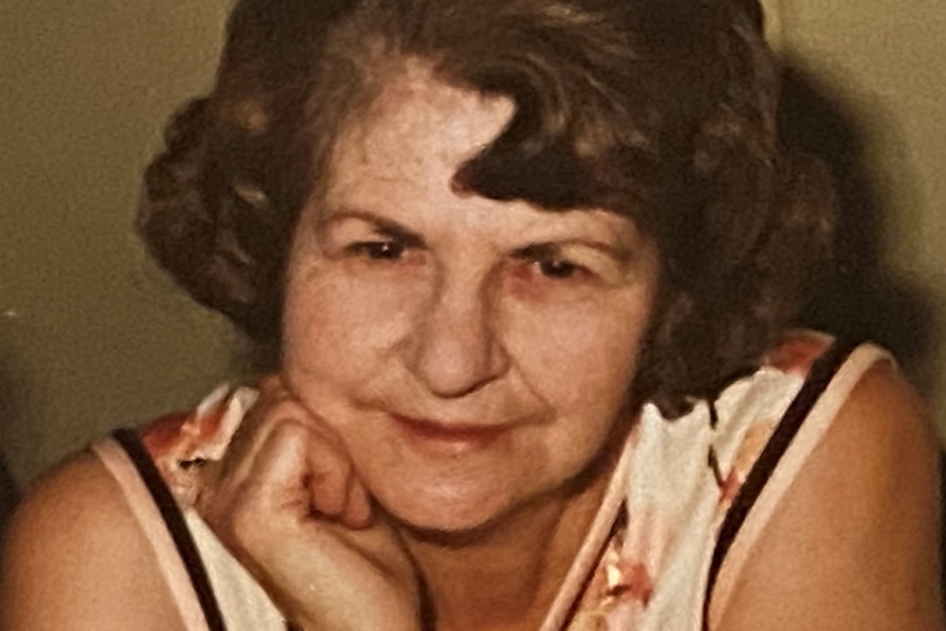 A grainy shot of a woman with a short dark brown bob, smiling with hand to her chin.