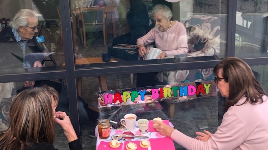 Four people celebrate a birthday - two are behind the window of an aged care home.