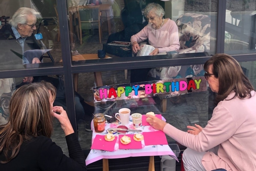 Four people celebrate a birthday - two are behind the window of an aged care home.