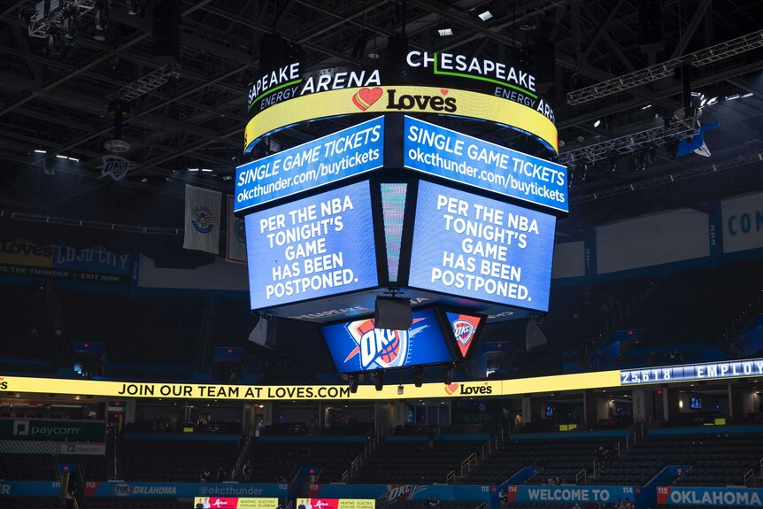 A big screen in the middle of a basketball stadium shows a sign saying 'per the NBA tonight's game has been postponed'