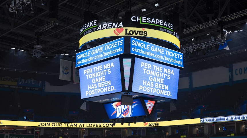 A big screen in the middle of a basketball stadium shows a sign saying 'per the NBA tonight's game has been postponed'