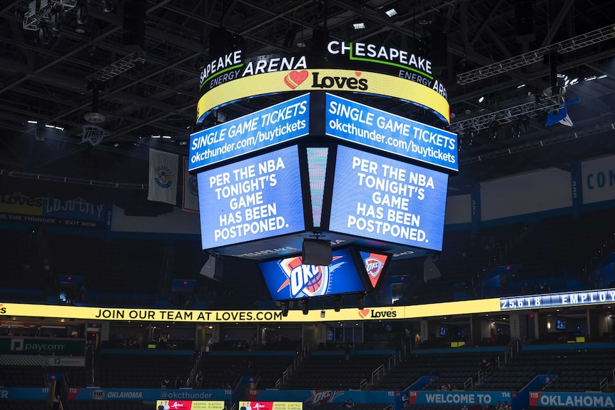 A big screen in the middle of a basketball stadium shows a sign saying 'per the NBA tonight's game has been postponed'