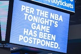 A big screen in the middle of a basketball stadium shows a sign saying 'per the NBA tonight's game has been postponed'