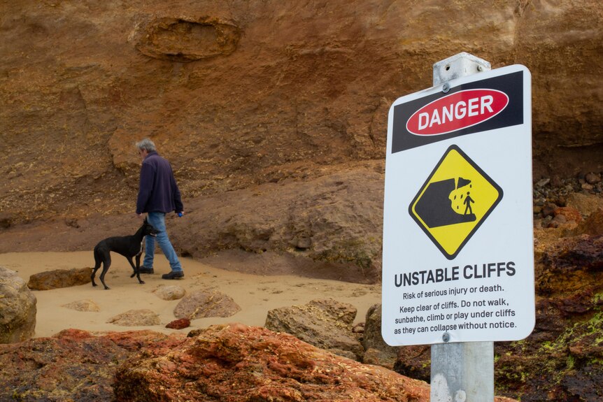 A man and his dog walk under cliffs with a sign next to them warning people to stay away from the cliffs due to erosion.
