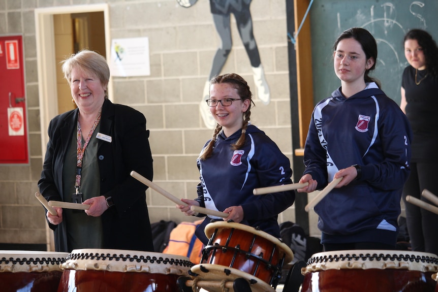 Two students in school uniform listen attentively as they hold drumsticks. They're joined by Alderman Beth Warren, smiling.