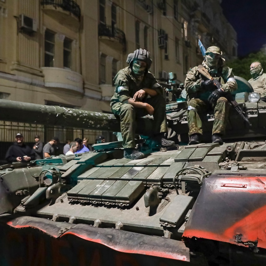 Three men in military uniforms and face masks sit on a tank on Russian street at night. The tank bears the letter Z in red.