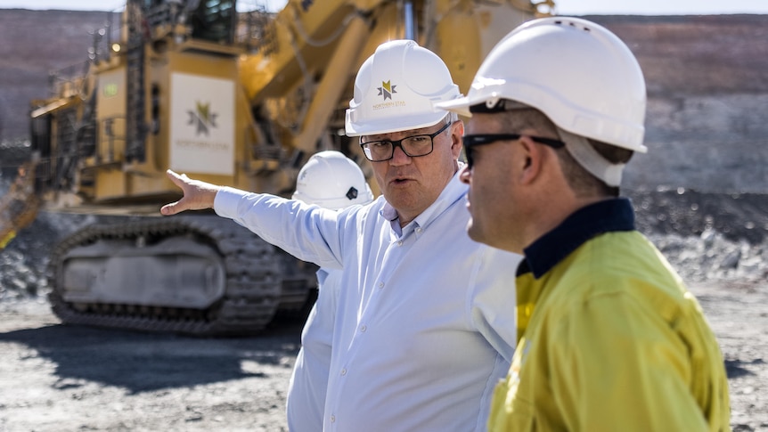 Prime Minister Scott Morrison pointing at mining machinery in the background while speaking to a mine worker wearing a hard hat.