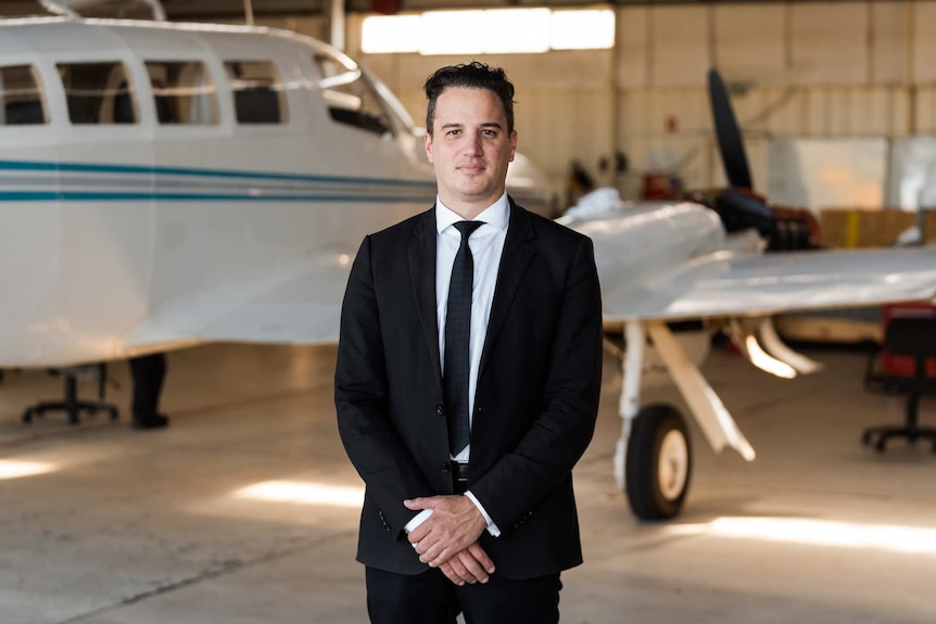 A man with short dark hair wearing a black suit and tie and white shirt stands in front of a small plane with his hands clasped.