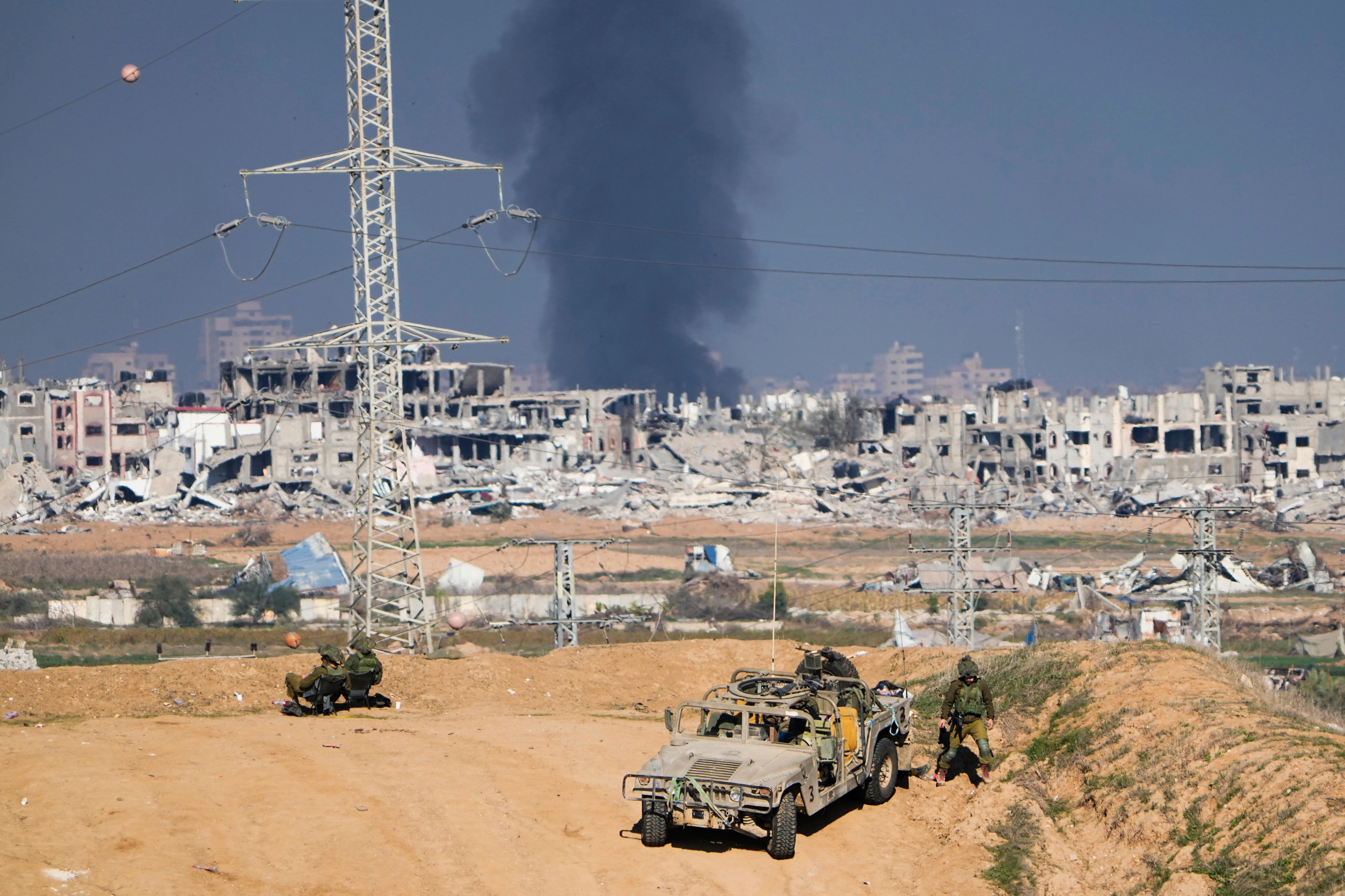 A military vehicle on a sandy embankment with troops standing and sitting nearby. Destroyed buildings and smoke in background