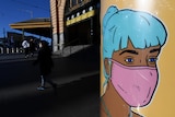 A close up of street art showing a woman with blue hair wearing a pink mask, with commuters walking in background