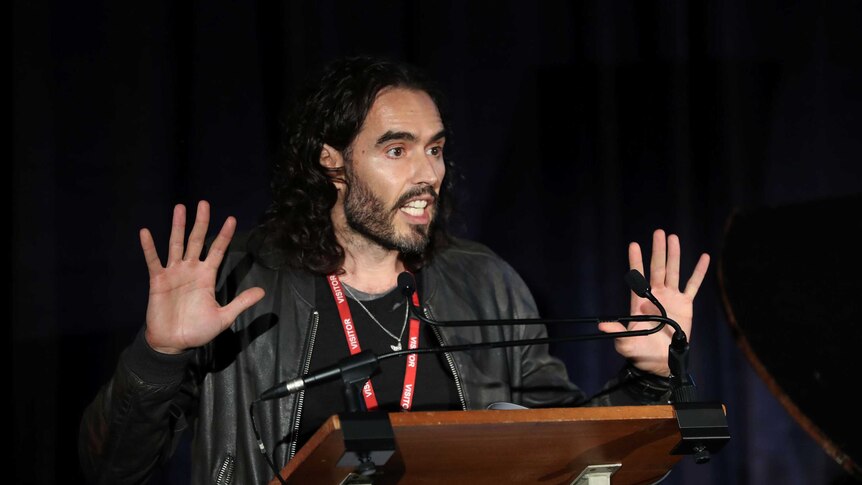 Russell Brand with raised hands stands behind a lectern looking intently ahead, in mid-speech.