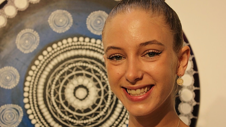 Close up image of Ursula Tate smiling in front of round blue dot painting in art gallery