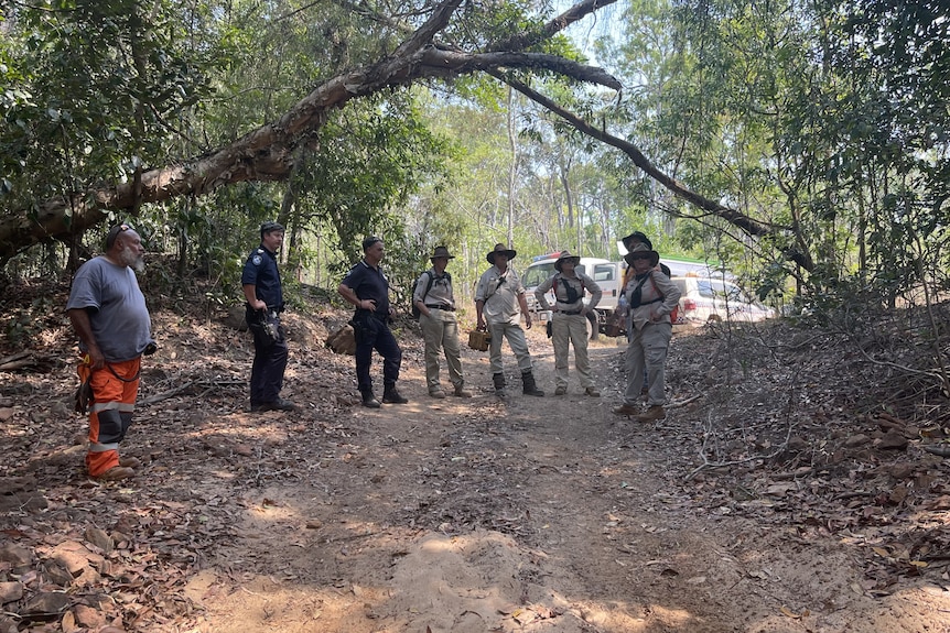 Personnel wearing uniforms stand on a dirt roadway in a forested area