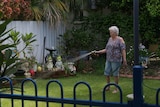 An elderly woman stands watering her garden.
