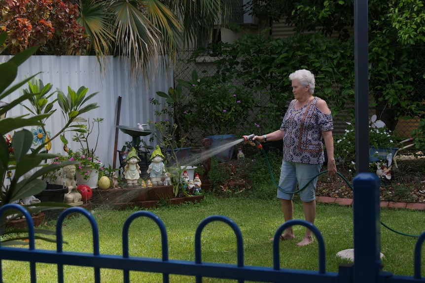 An elderly woman stands watering her garden.