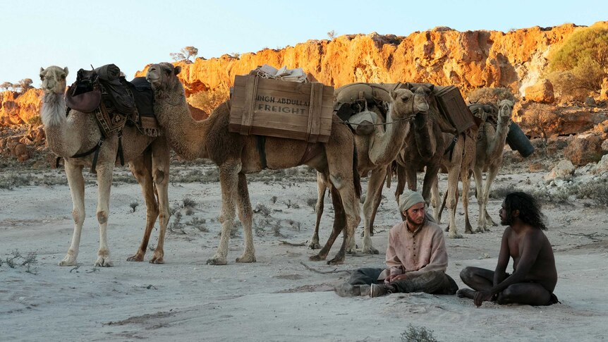 A group of camels loaded with freight boxes stand next to two male actors sitting cross legged in the sand