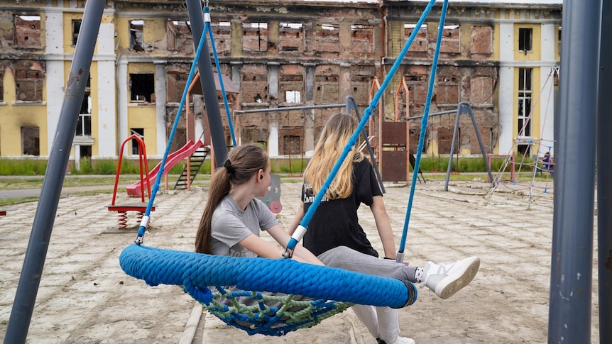Two teenage girls sit on a swing chair, looking towards the damaged school building