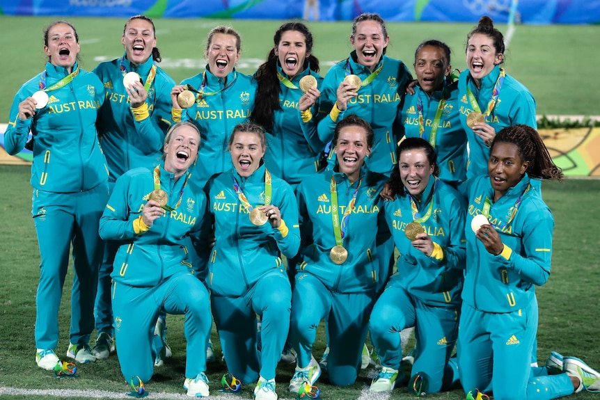 A team of women's rugby players in Australian tracksuits pose with medals.