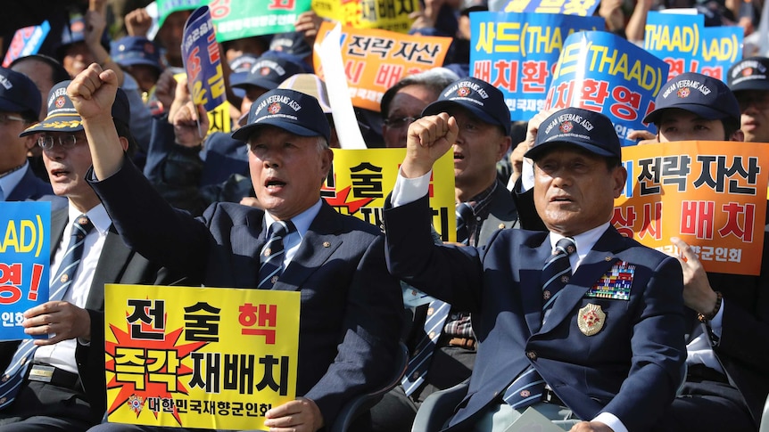 Men in uniform chant with their hands in the air as they hold protest signs in Korean language.