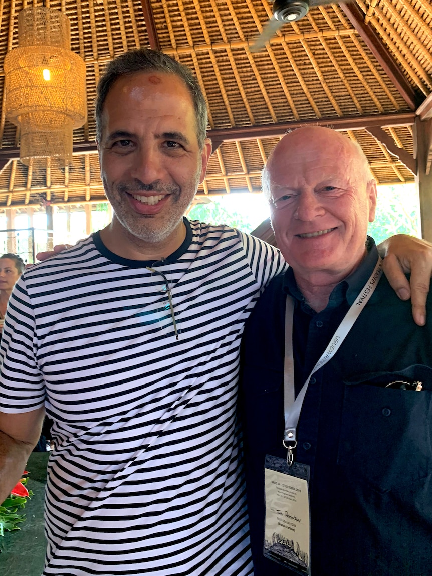 Two men with arms on each other's shoulders smile beneath a thatched roof