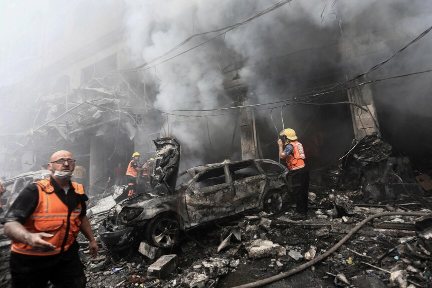 Three Palestinian men in orange hi-vis vests stand beside and on the smoking wreckage of a building and car.