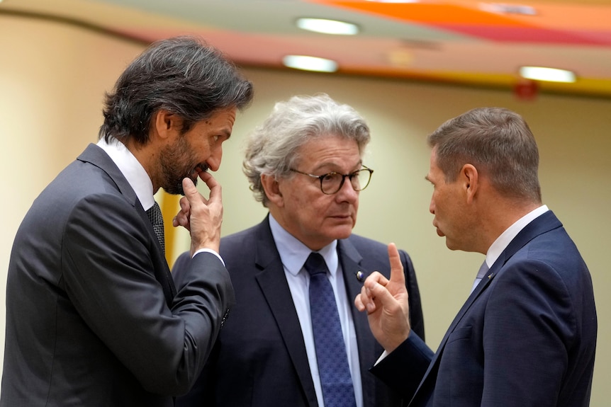 Three men in suits are pictured talking to each other, two are pictured from the side and one from the front. 