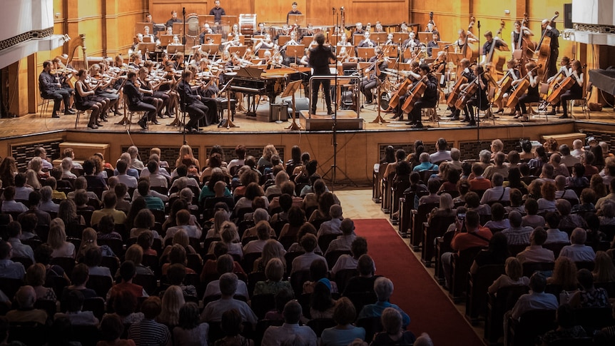 A view from the back of a large concert hall showing a full audience and an orchestra on stage.