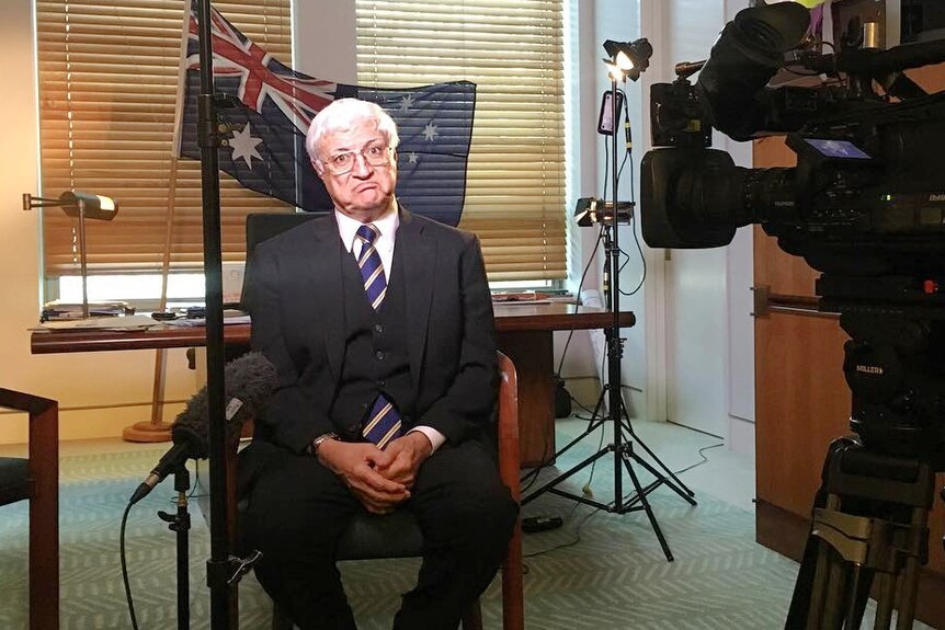 Bob Katter in suit sits in office about to do a media interview, pulling a funny face at the camera