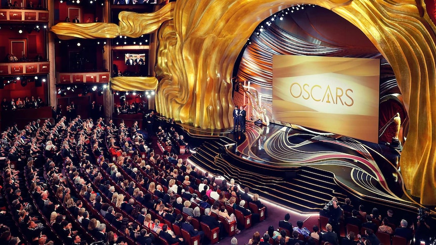 A large theatre filled with people watching presenters on stage in front of a large screen with "Oscars" displayed on it.