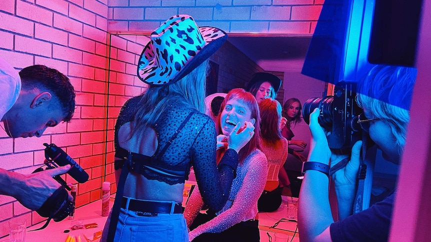 A cramped bathroom scene lit in pink and purple, a woman wearing a cowgirl hat does the makeup of another.