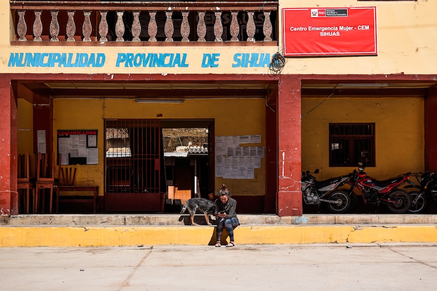 A woman and a dog sit on a concrete step in front of a shop. 