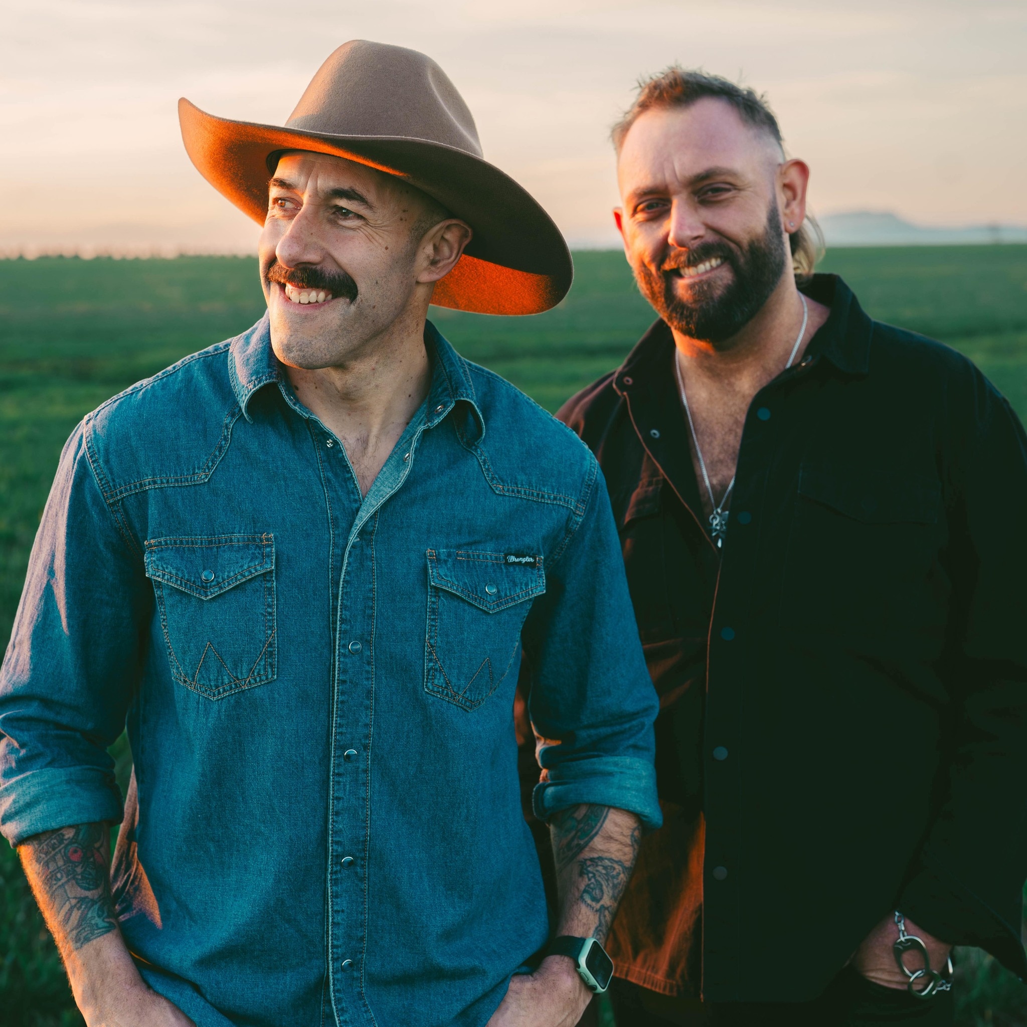 dois homens sorrindo em um campo, um com chapéu de cowboy e o outro com barba curta.