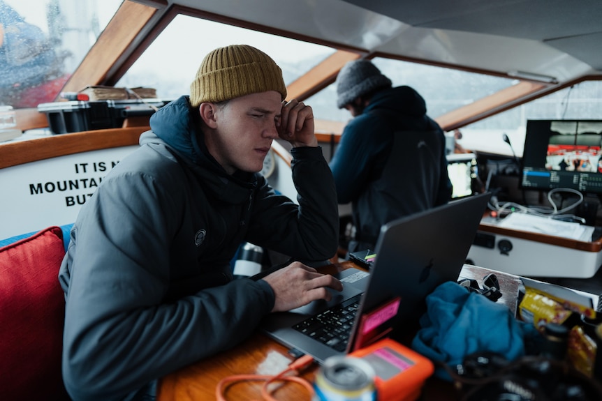 A young man sitting inside a small boat works at a laptop editing photos.