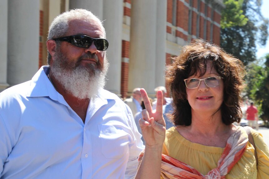 A man and a woman who is making a peace sign stand outside court looking pleased.
