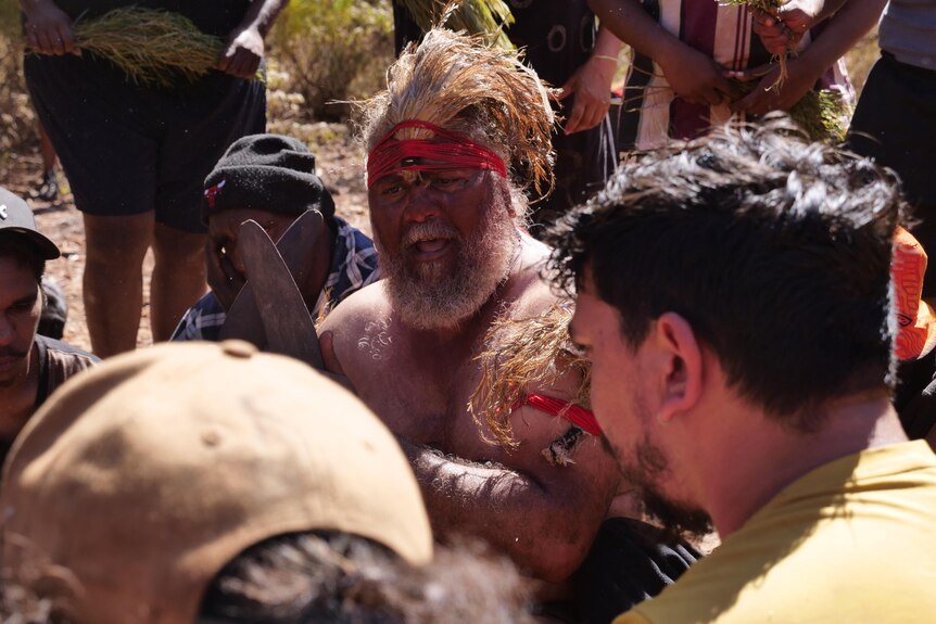 An indigenous man in traditional dress delivers a performance as a crowd watches on.