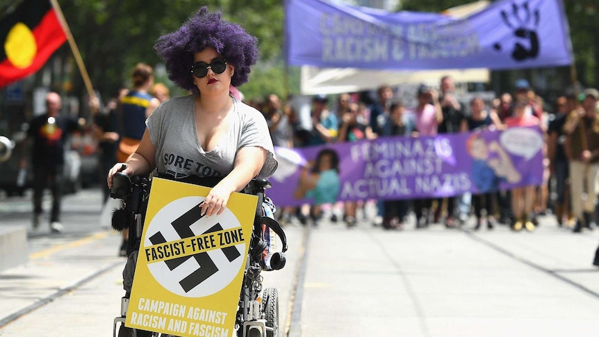 A woman with purple hair holds a sign saying 'Fascist free zone' with protesters in the background.