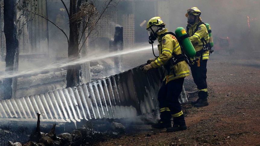 Two firefighters dressed in yellow protective clothing spray a burnt out building.