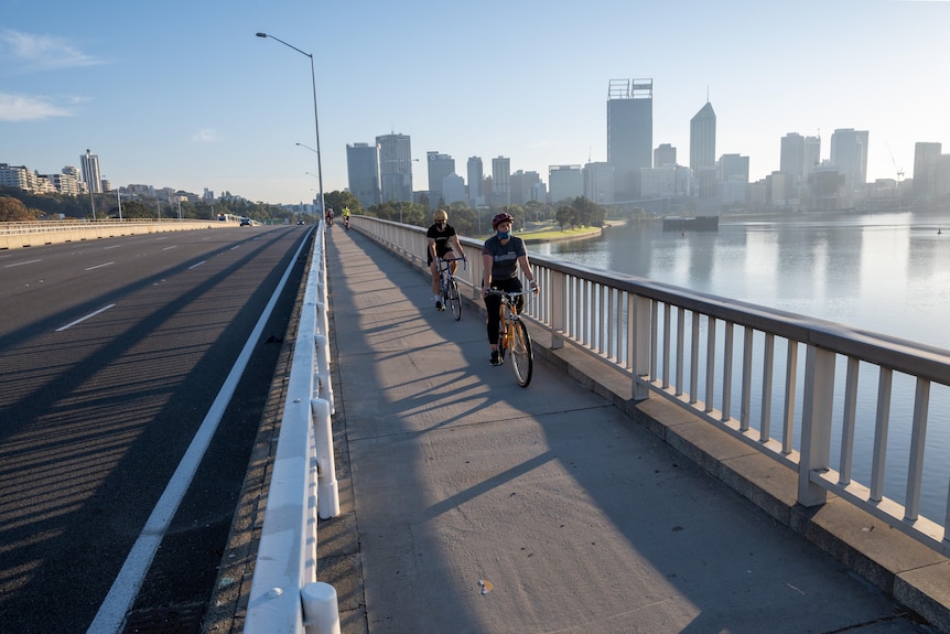 Two cyclists wearing masks ride across bridge with Perth city skyline in background. 