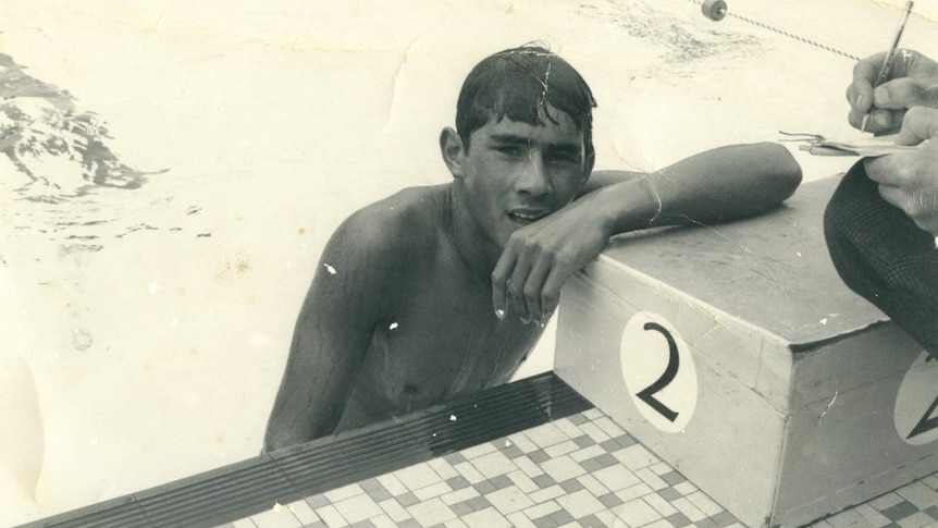 A young man half-immersed in water leans on the dicing block at the end of a pool lane.