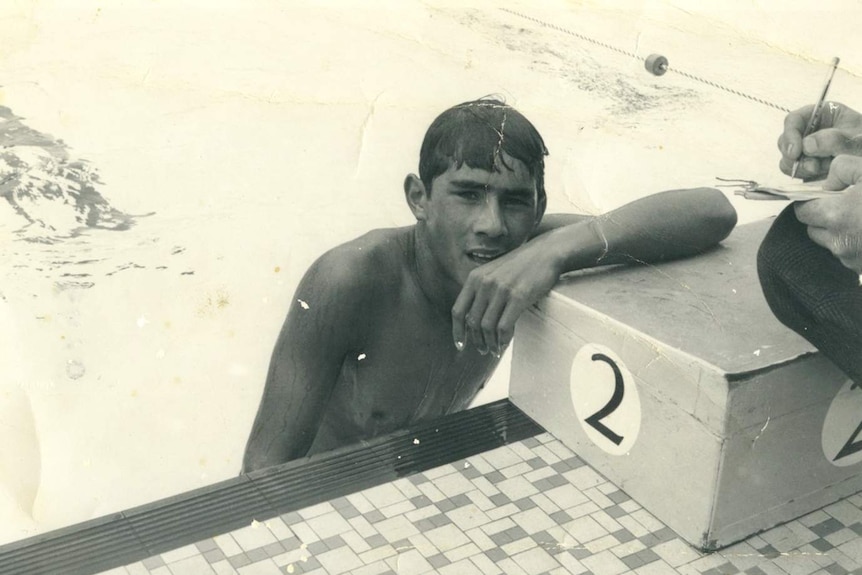 A young man half-immersed in water leans on the dicing block at the end of a pool lane.