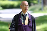 Bald man wearing navy blue robes, holding stick, and walking in green outdoors.