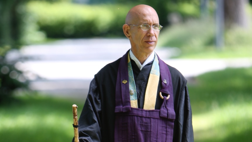 Bald man wearing navy blue robes, holding stick, and walking in green outdoors.